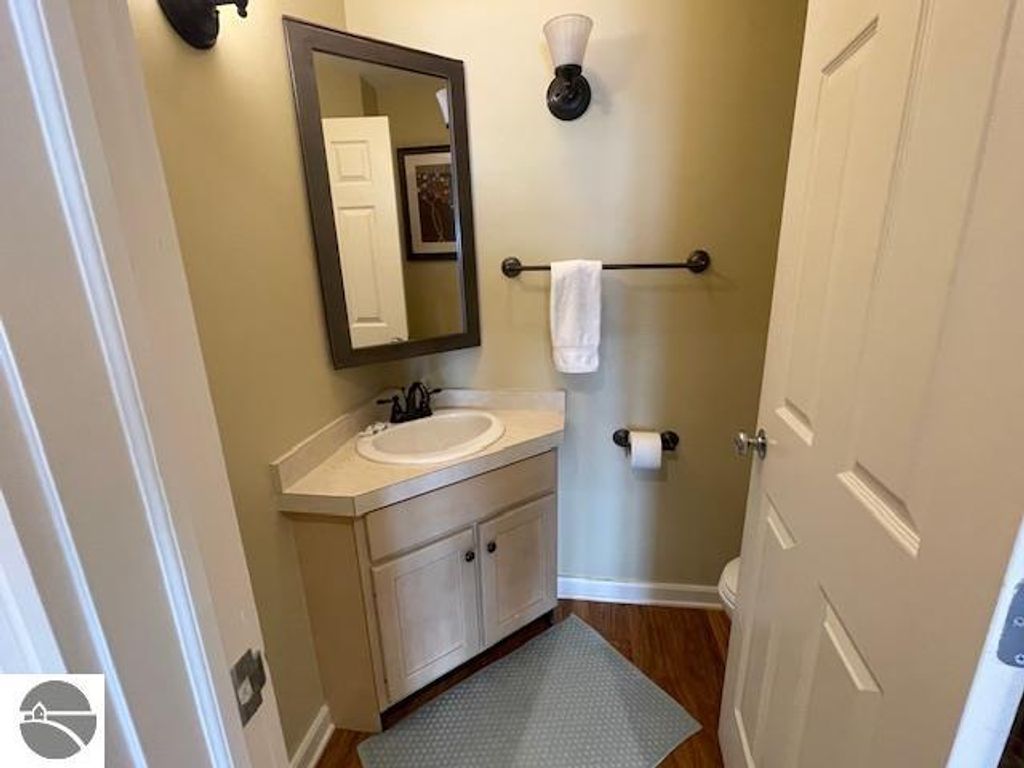Compact bathroom with a sink, mirror, and towel rack, featuring neutral walls and wooden flooring, highlighting the updated furnishings of the 12C Pinnacle Place listing in Glen Arbor, MI.