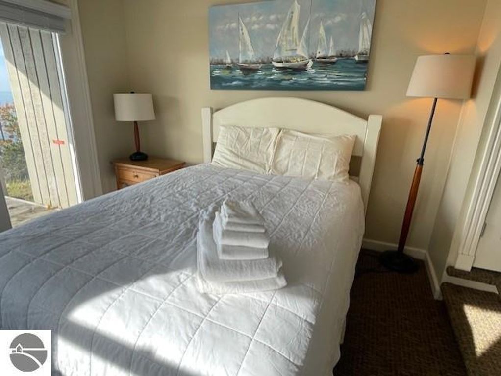 Cozy bedroom with a white quilted bedspread, decorative sailing artwork, and bedside lamps, featuring natural light and a view of Lake Michigan.