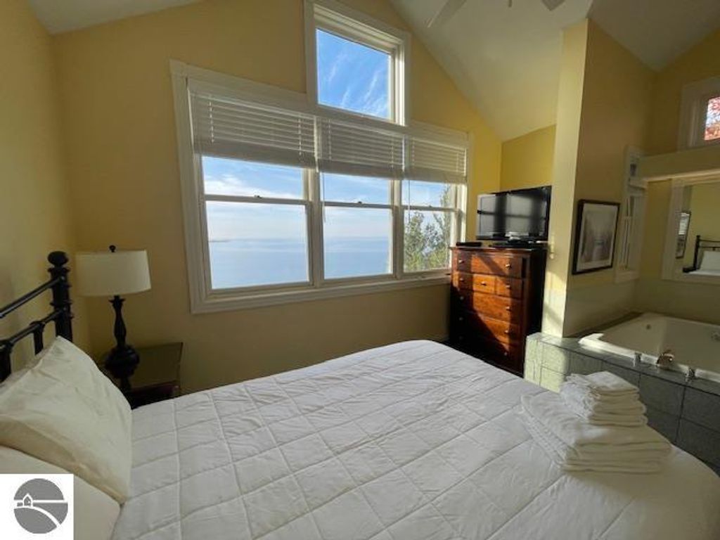 Bright bedroom with a queen bed, dresser, and television, featuring large windows showcasing panoramic views of Lake Michigan and surrounding landscape.