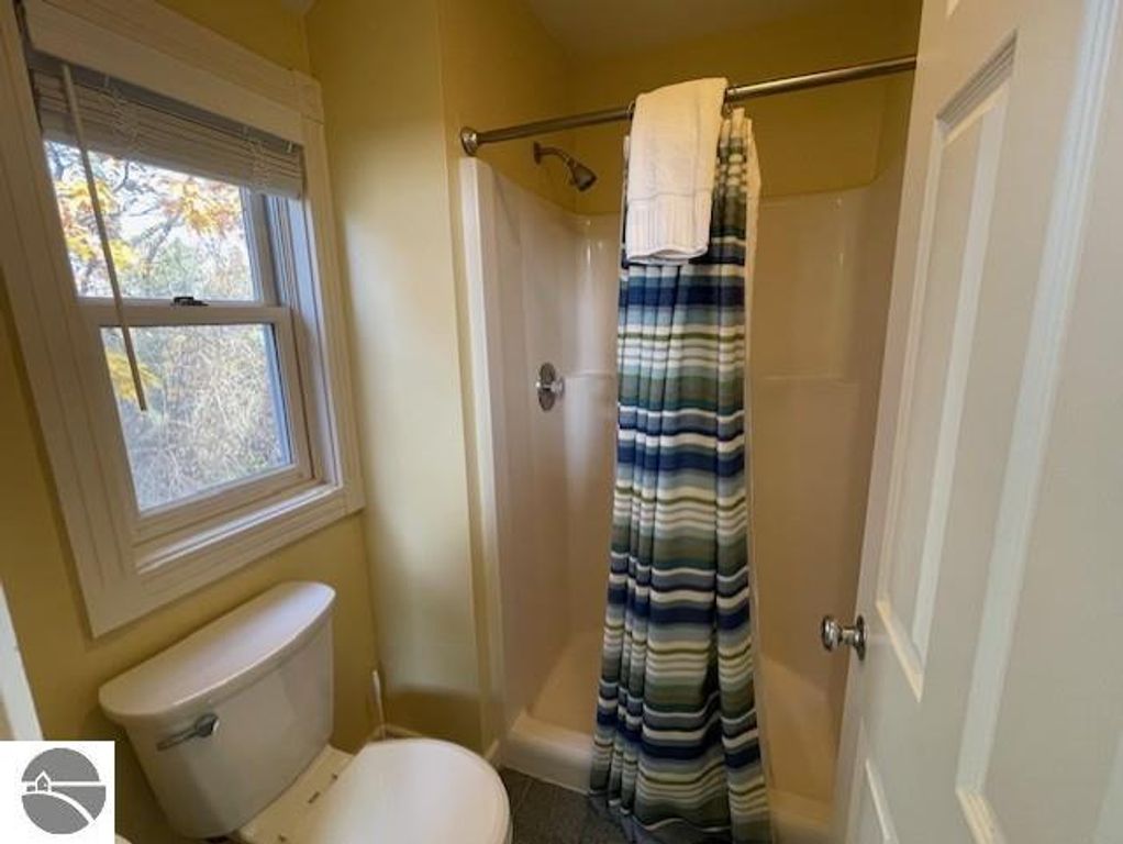 Bathroom interior featuring a shower with a striped curtain, toilet, and window allowing natural light, showcasing the updated amenities of the 12C Pinnacle Place unit in Glen Arbor, MI.