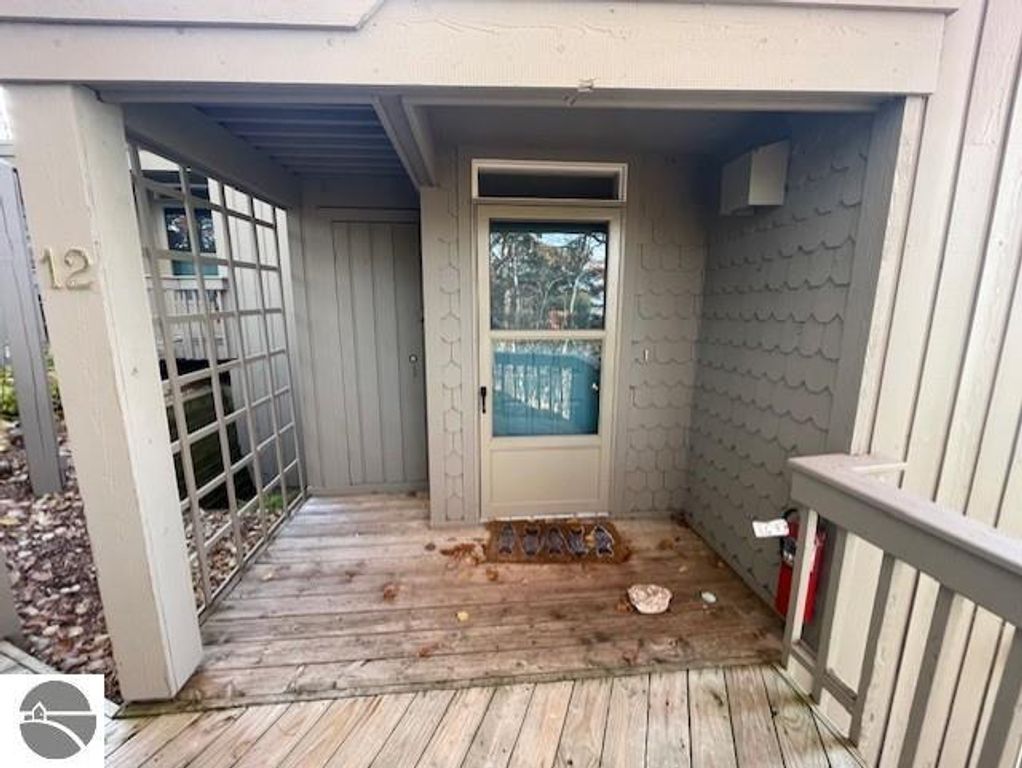 Entrance to 12C Pinnacle Place, Glen Arbor, featuring a wooden deck, door with window, and nearby lattice structure, showcasing the property&rsquo;s access point in a resort setting.
