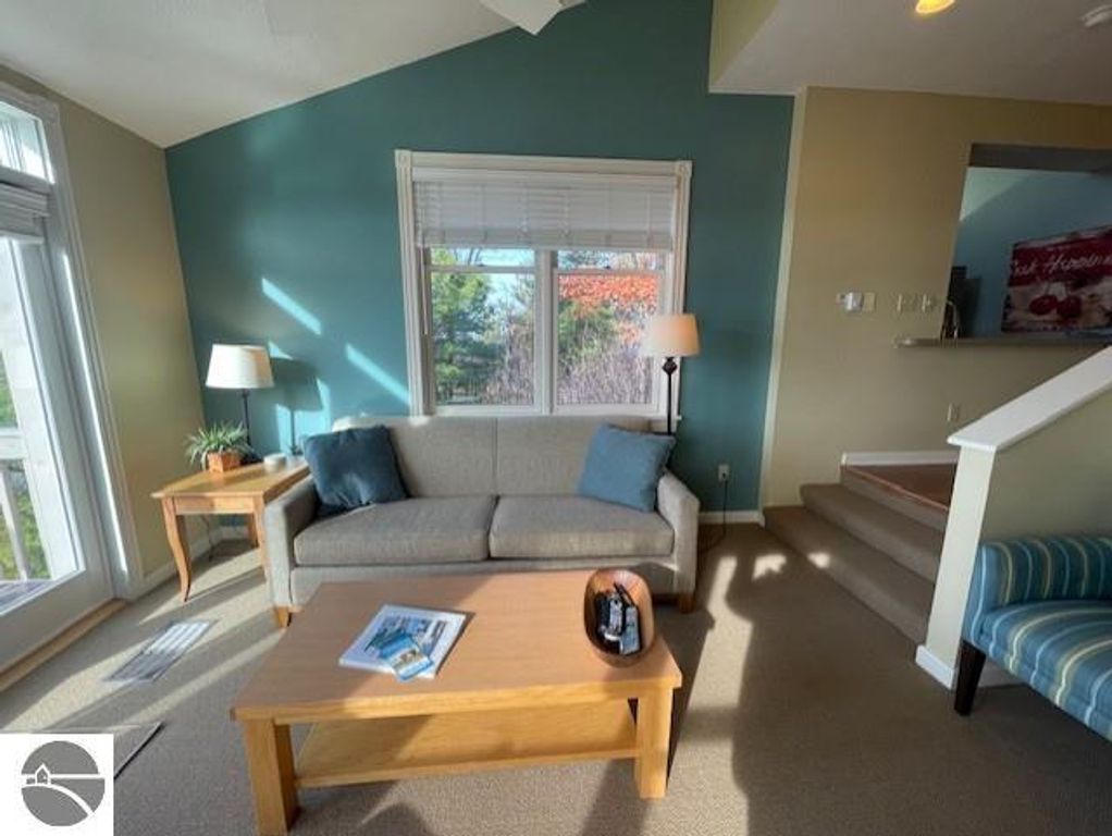 Living room interior of 12C Pinnacle Place, featuring a gray sofa with blue cushions, wooden coffee table, lamp, and staircase, showcasing a cozy atmosphere with natural light and views of Lake Michigan.