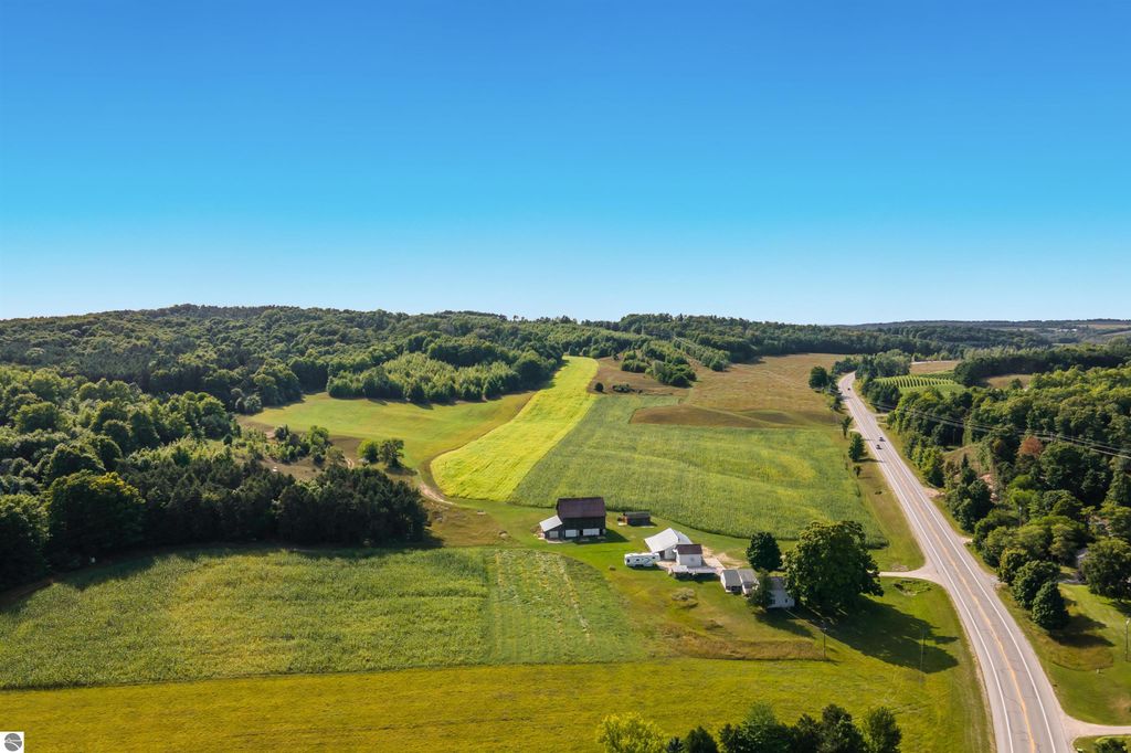 Aerial view of 74.2 acres of farmland in Suttons Bay, Michigan, featuring rolling green fields, wooded areas, and nearby road, highlighting potential development opportunities and scenic landscapes.