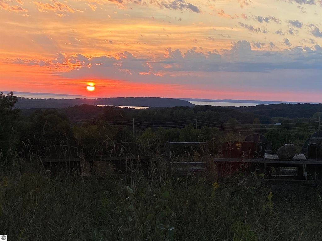 Sunset over Lake Michigan and Grand Traverse Bay, with vibrant orange and pink hues, framed by lush greenery and rustic outdoor seating in the foreground, highlighting the scenic beauty of Suttons Bay, MI.