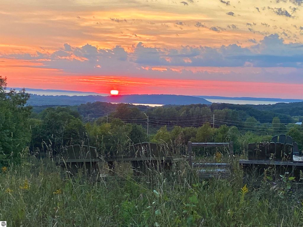 Sunset view over lush greenery and distant water, showcasing vibrant orange and pink skies, with silhouettes of chairs in the foreground, highlighting the scenic beauty of Suttons Bay, MI.