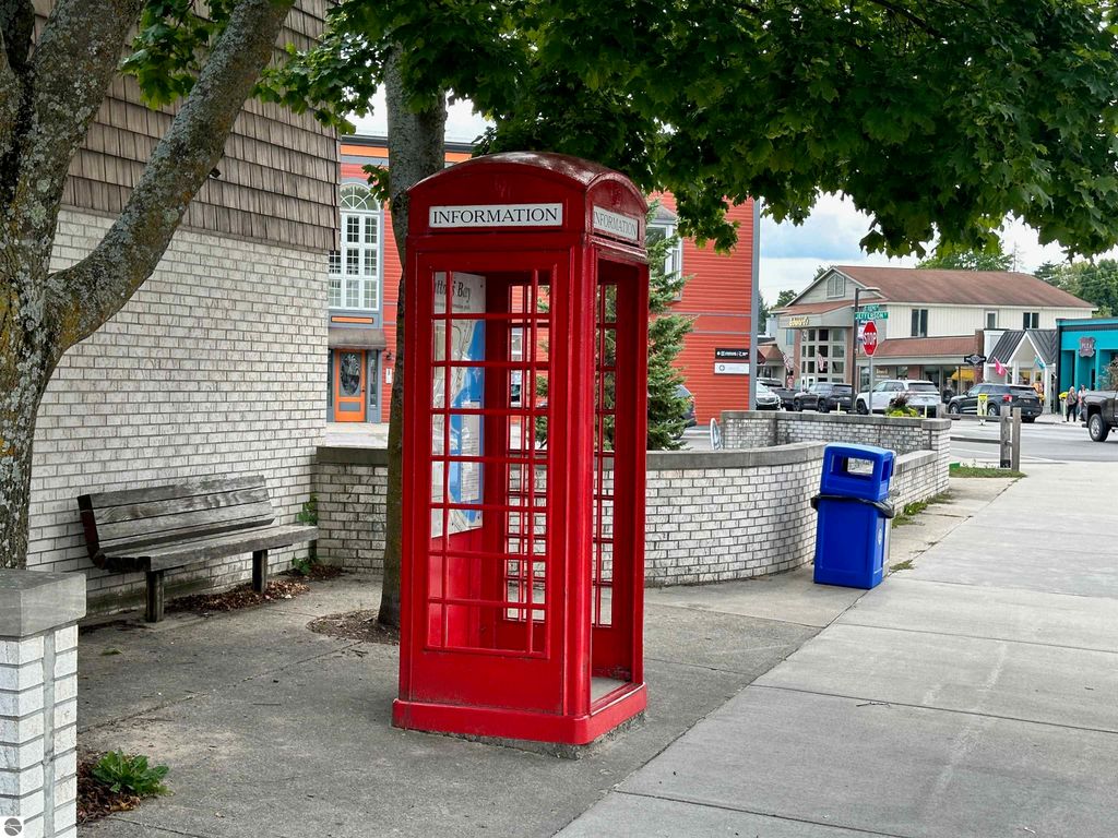 Red information phone booth on sidewalk in Suttons Bay, MI, near a bench and colorful storefronts, providing local visitor information.