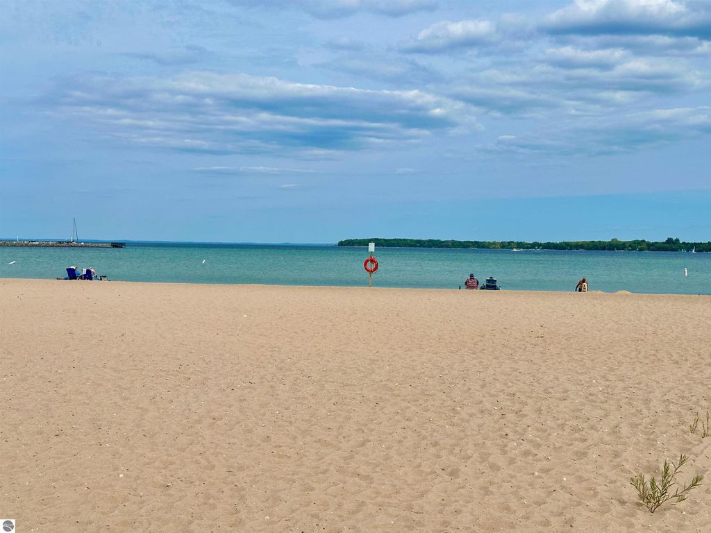 Beach scene at Suttons Bay, MI, featuring sandy shore, calm waters, and distant boats, with two people seated on beach chairs enjoying the view.