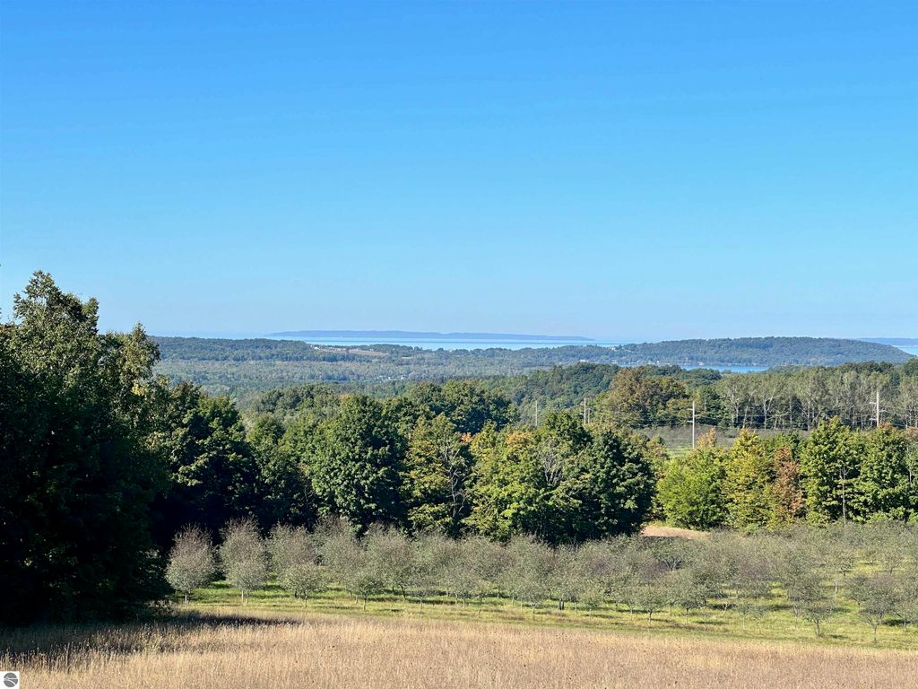 Scenic view of high-elevation farmland in Suttons Bay, showcasing lush greenery, distant water views of Grand Traverse Bay, and rolling hills under a clear blue sky, highlighting the parcel's development potential and breathtaking landscapes.