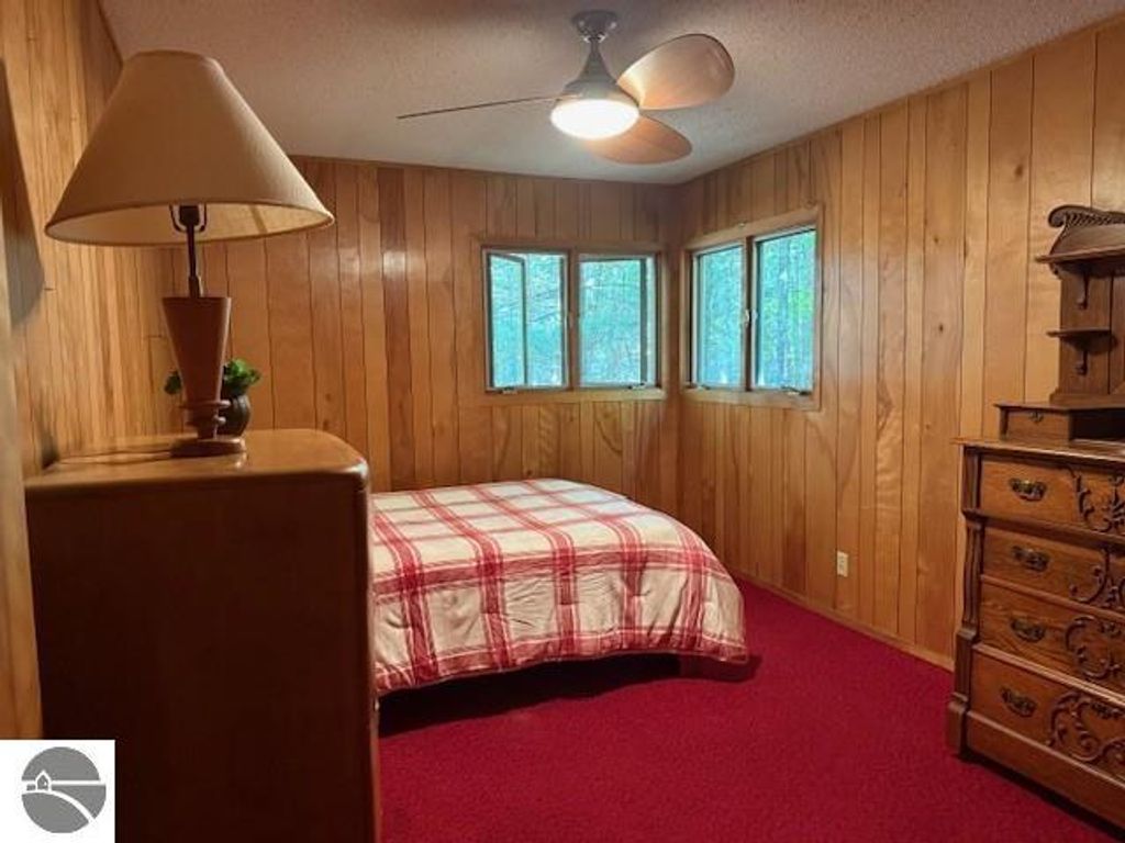 Cozy bedroom with wooden paneling, red carpet, plaid bedding, bedside lamp, and windows overlooking wooded area, showcasing the inviting atmosphere of the Glen Arbor cottage listed for sale.