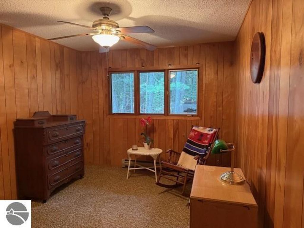 Cozy interior of a cottage at 6025 Forest Haven Drive, Glen Arbor, featuring wooden paneling, a ceiling fan, windows overlooking greenery, a rocking chair, and vintage furniture, highlighting the property's tranquil Up North atmosphere.