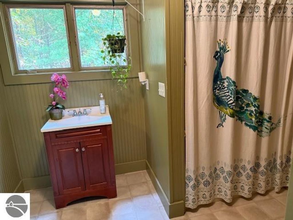 Cozy bathroom featuring a wooden vanity with sink, pink orchid plant, and a peacock-patterned shower curtain, surrounded by natural light from windows in a serene setting.