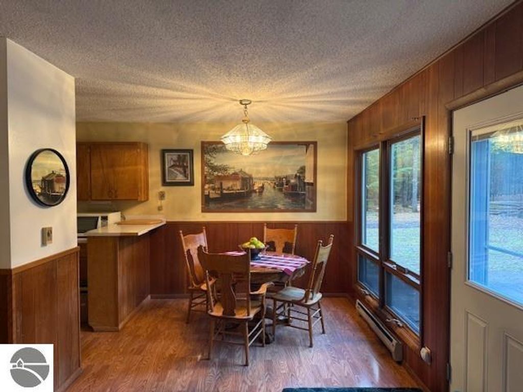 Cozy dining area with wooden table and chairs, featuring a framed painting and large windows overlooking wooded surroundings, in a cottage at 6025 Forest Haven Drive, Glen Arbor, MI.