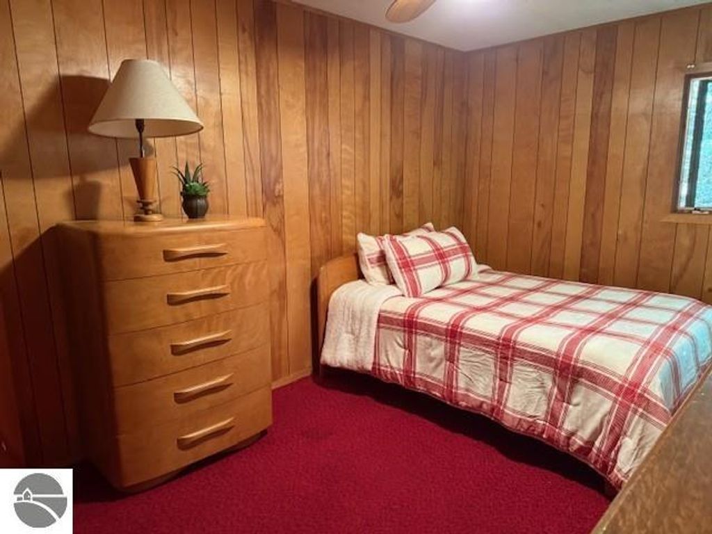 Cozy bedroom featuring a twin bed with red and white plaid bedding, wooden panel walls, a stylish wooden dresser with five drawers, a bedside lamp, and a small potted plant, reflecting the inviting atmosphere of the Glen Arbor cottage for sale.