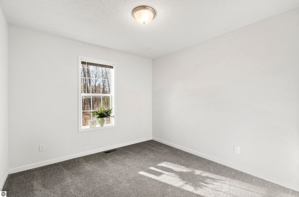 Bright and airy interior of a new bedroom in a ranch-style home, featuring a window with a view of trees, soft gray carpet, and neutral walls, showcasing the inviting atmosphere of the property at 11754 Country Acres Drive, Interlochen, MI.