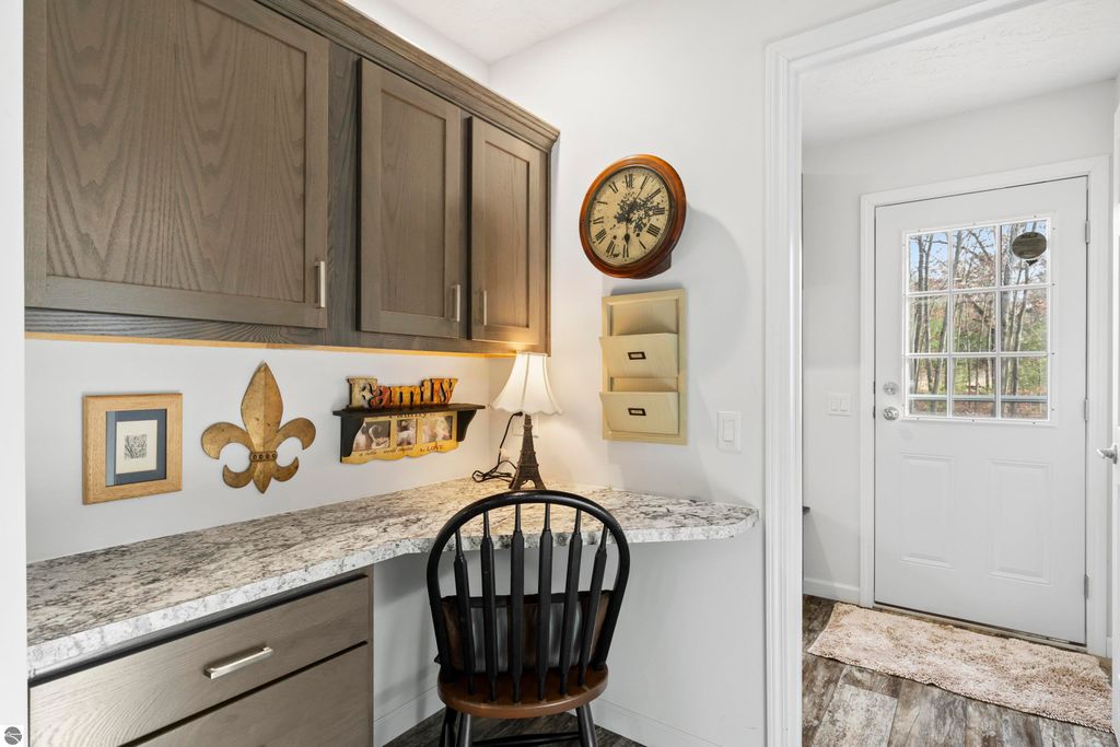 Home office nook with dark cabinetry, granite countertop, decorative items including a family sign and clock, and a door leading outside in a newly built ranch home in Interlochen, MI.