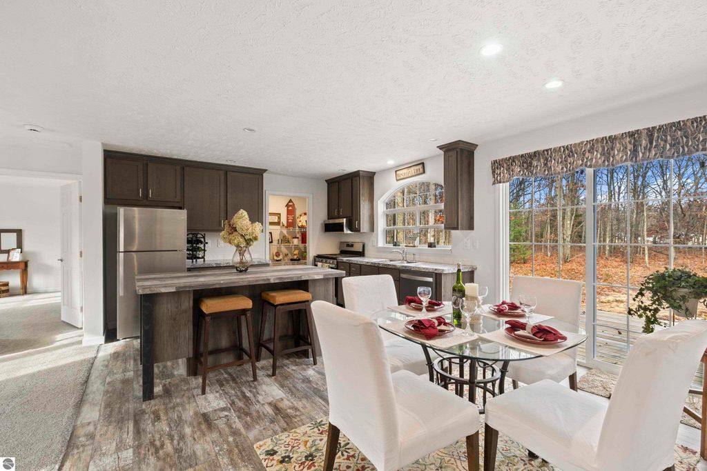 Modern kitchen and dining area featuring a table set for four, stainless steel appliances, dark cabinetry, and large windows overlooking a wooded backyard in a newly listed home at 11754 Country Acres Drive, Interlochen, MI.