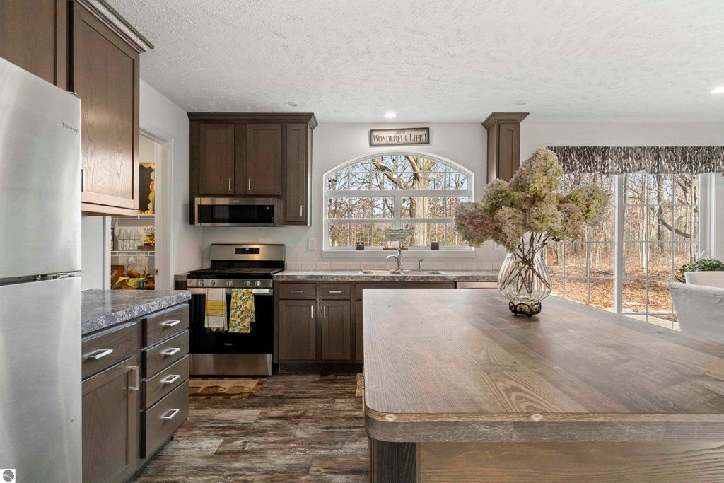 Modern kitchen interior with stainless steel appliances, dark wood cabinets, and a large wooden island, featuring a vase of flowers and large windows overlooking a wooded area, highlighting the home's inviting atmosphere in Interlochen, MI.