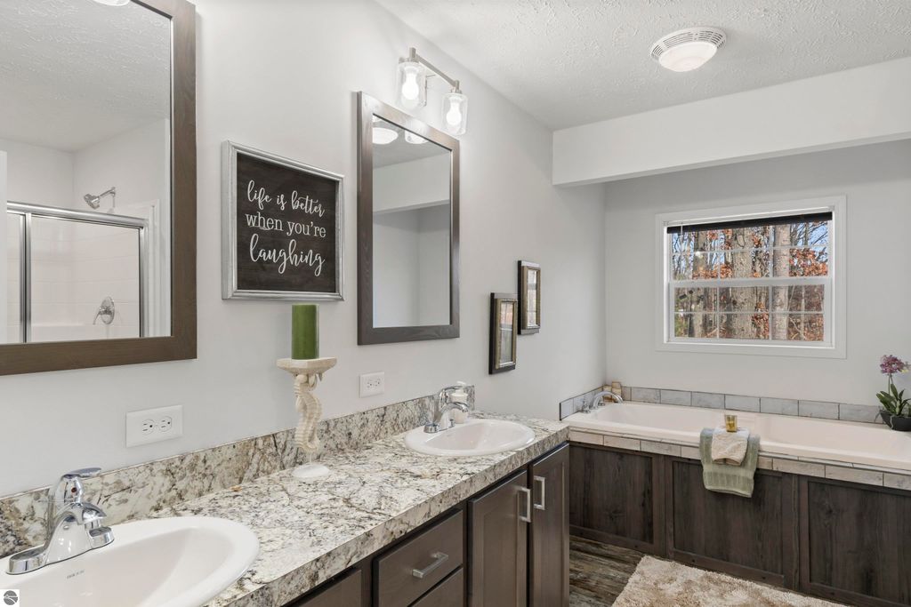 Modern bathroom with dual sinks, granite countertops, framed mirrors, decorative wall sign, and a bathtub, showcasing a spacious and inviting design in a new home at 11754 Country Acres Drive, Interlochen, MI.