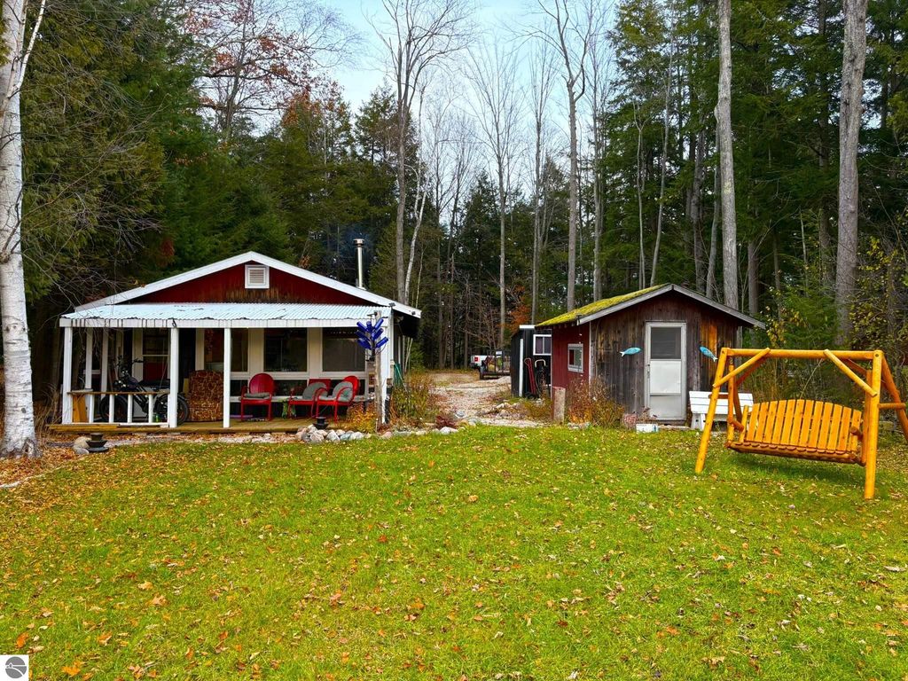 Waterfront cottage at 2456 Birch Road, Grawn, MI, featuring a red exterior, porch with seating, grassy yard, and additional outbuildings, surrounded by trees.