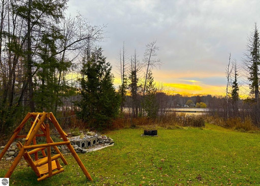 Lakefront view from the backyard of a cottage in Grawn, MI, featuring a wooden swing set, fire pit, and surrounding trees, with a sunset illuminating the sky over Tonawanda Lake.