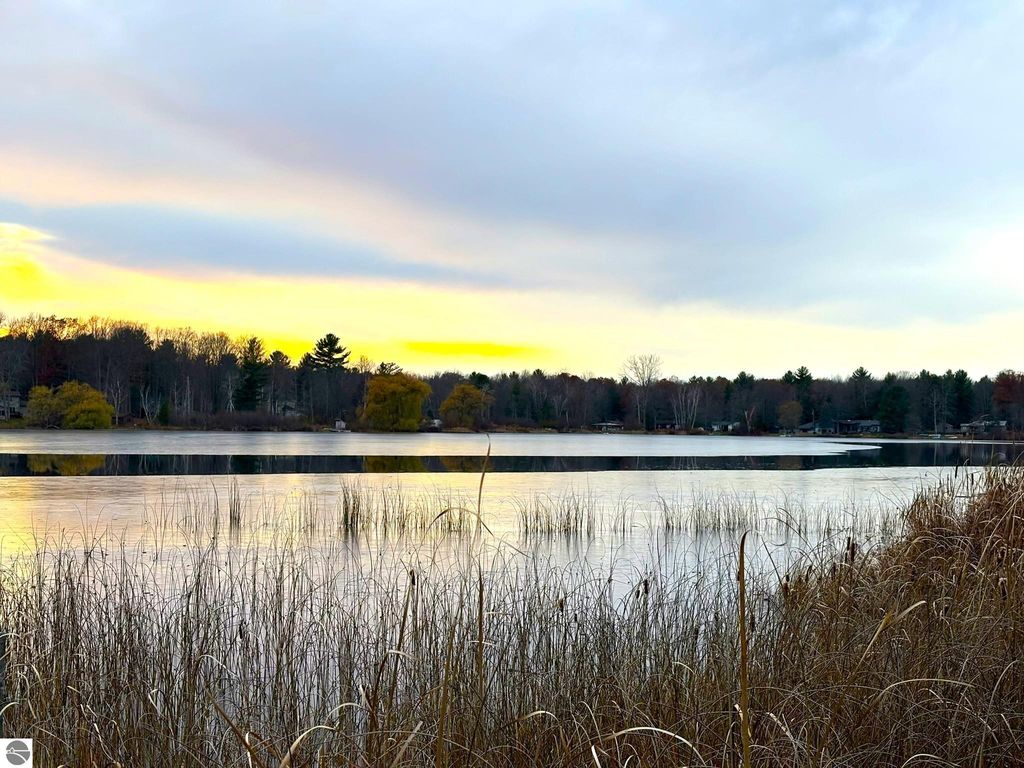 Scenic view of Tonawanda Lake at sunset, reflecting clouds and trees, with tall grasses in the foreground, highlighting the peaceful waterfront setting near the cottage for sale at 2456 Birch Road, Grawn, MI.