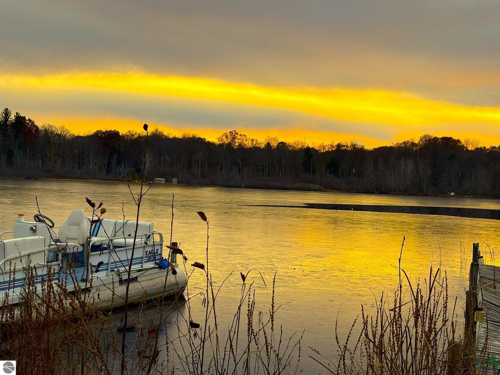 Waterfront view of Tonawanda Lake at sunset, featuring a pontoon boat moored near the shore, surrounded by tall grasses and trees, highlighting the serene natural landscape ideal for year-round living.