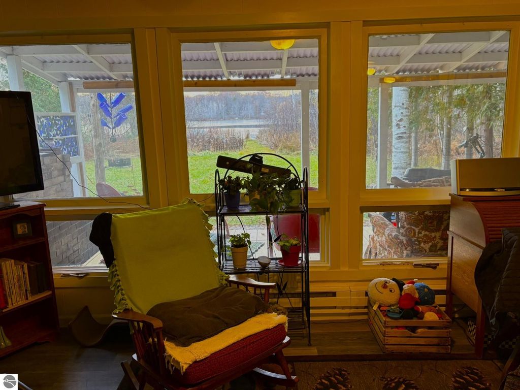 Cozy interior view of a waterfront cottage at 2456 Birch Road, featuring a rocking chair, potted plants, and large windows overlooking a serene outdoor setting by Tonawanda Lake.