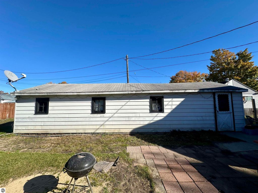 Back view of a bungalow in Traverse City, featuring a satellite dish, fenced yard, and patio area with a grill, showcasing the exterior condition and surroundings.