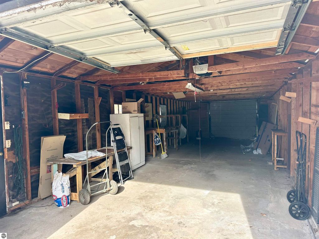 Garage interior with open door, featuring wooden beams, storage shelves, and various items including boxes and a folding chair, highlighting additional storage space for the bungalow at 224 W Fifteenth Street, Traverse City, MI.