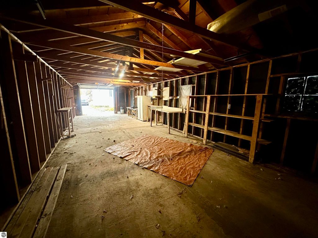 Interior of a spacious garage with exposed wooden beams, dirt floor, and shelves along the walls, featuring a tarp on the ground and natural light coming from an open door.