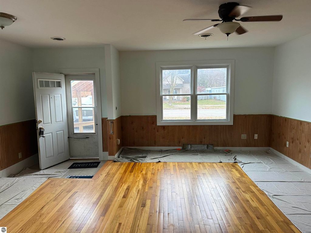 Interior view of a freshly painted bungalow living room at 224 W Fifteenth Street, Traverse City, featuring hardwood floors, a ceiling fan, and large windows, with a welcoming entrance and partially covered floor.