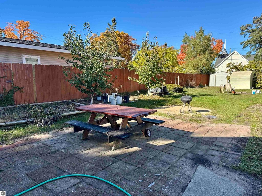 Backyard of a bungalow in Traverse City, featuring a picnic table, grill, fenced area, and garden space with trees, showcasing autumn foliage and nearby storage shed.