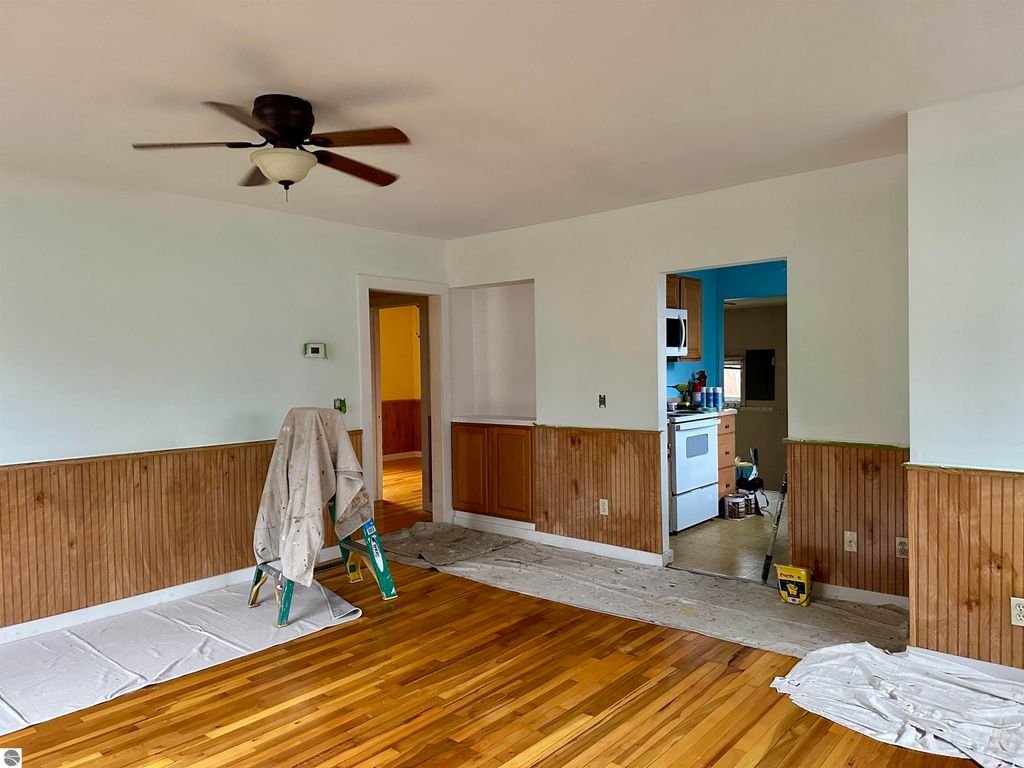 Living room interior of a bungalow in Traverse City, featuring hardwood floors, a ceiling fan, and freshly painted walls, with a view into the kitchen area.
