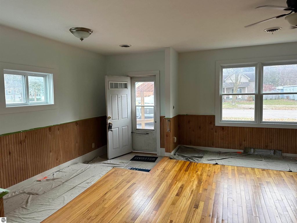 Interior view of a freshly painted bungalow living room in Traverse City, featuring hardwood floors, a front door, and large windows, with a partially finished look and protective coverings on the floor.