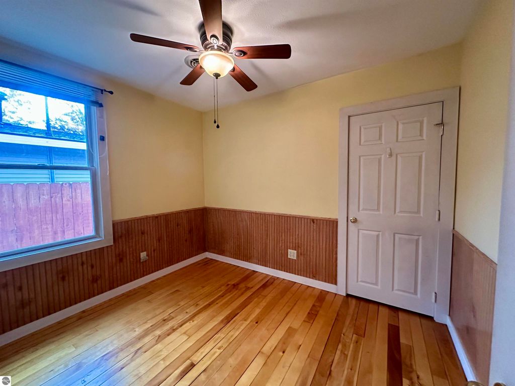 Cozy bedroom with hardwood floors, ceiling fan, and window, featuring yellow walls and wainscoting, located in a bungalow in downtown Traverse City.