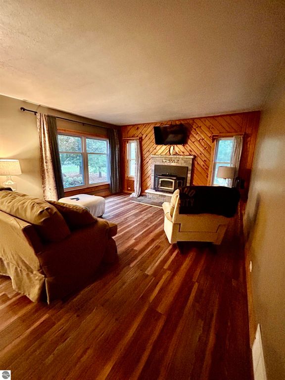 Cozy living room featuring a plush sofa and armchair, wooden flooring, large windows with curtains, and a stone fireplace with a mounted TV, showcasing a warm and inviting atmosphere ideal for family gatherings at 1007 W First Street, Tawas City, MI.