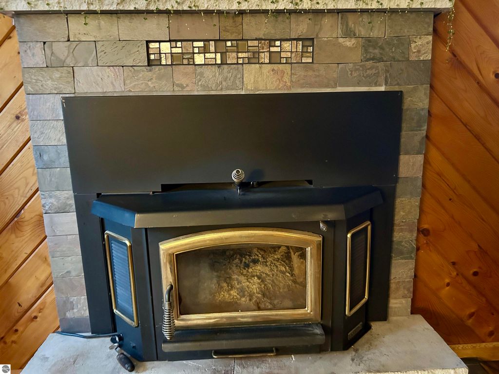 Wood-burning stove with ornate black and gold design, set against a stone and wood-paneled fireplace backdrop, highlighting cozy living space features in Tawas City home listing.
