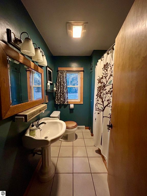 Bathroom featuring a pedestal sink, wooden accents, decorative shower curtain with tree design, and natural light from window, reflecting a cozy, inviting atmosphere in the Tawas City home listing.