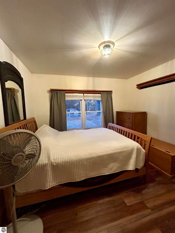 Cozy bedroom featuring a wooden bed with a white blanket, a standing fan, and natural light from a window with gray curtains, highlighting a peaceful retreat in Tawas City, MI.