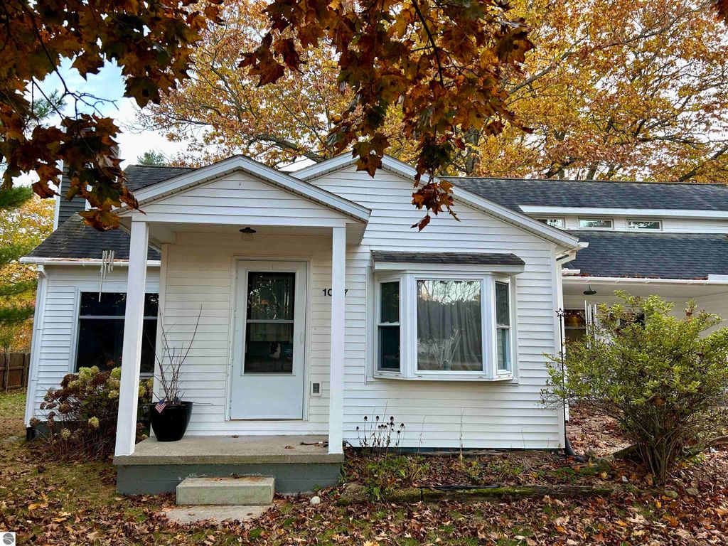 Charming white home at 1007 W First Street, Tawas City, MI, featuring a welcoming porch, surrounded by autumn foliage and greenery, ideal for families seeking tranquility and convenience.