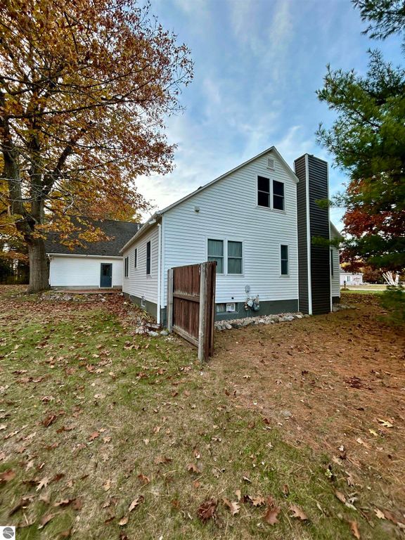 Exterior view of a residential home at 1007 W First Street, Tawas City, MI, showcasing a white facade, sloped roof, and surrounded by autumn foliage, emphasizing a tranquil setting.