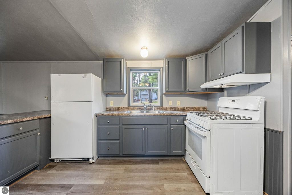Modern kitchen with gray cabinets, white refrigerator, and gas stove, featuring new flooring and natural light from a window, showcasing the interior of a move-in ready home for sale at 8401 Independence Avenue, Cadillac, MI.