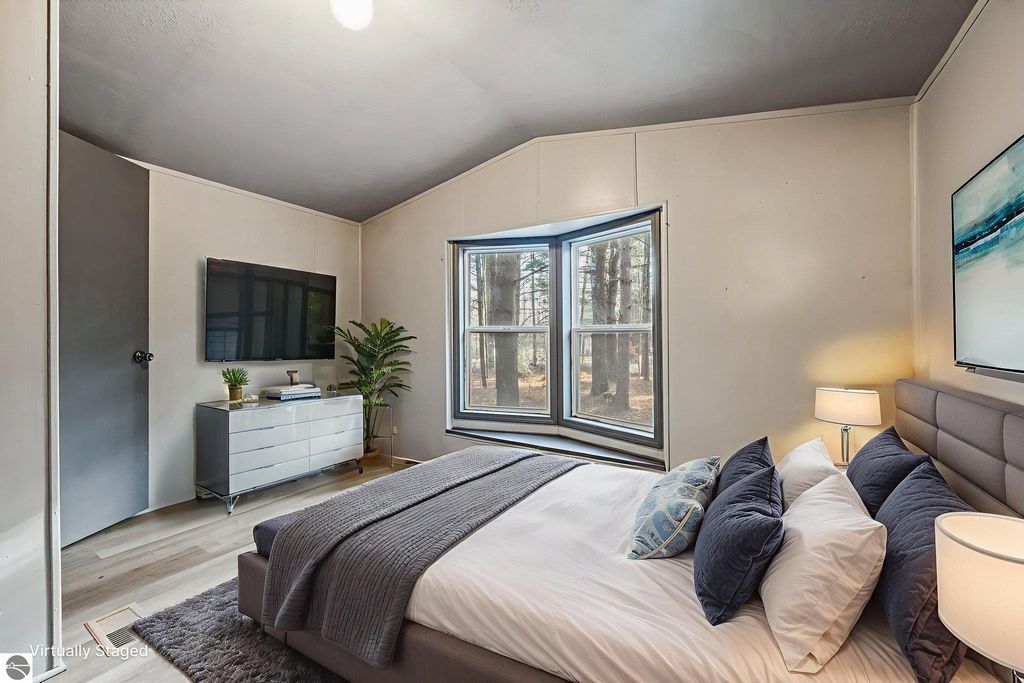 Cozy bedroom featuring a bed with decorative pillows, a modern dresser, a wall-mounted TV, and large windows overlooking a wooded area, showcasing a move-in ready home at 8401 Independence Avenue, Cadillac, MI.
