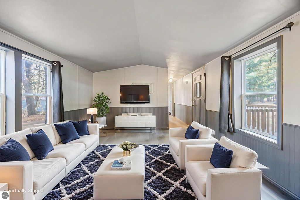 Modern living room featuring a white sofa with blue accent pillows, a large area rug, a television mounted on the wall, and large windows providing natural light, showcasing the cozy interior of the 3-bedroom home at 8401 Independence Avenue, Cadillac, MI.