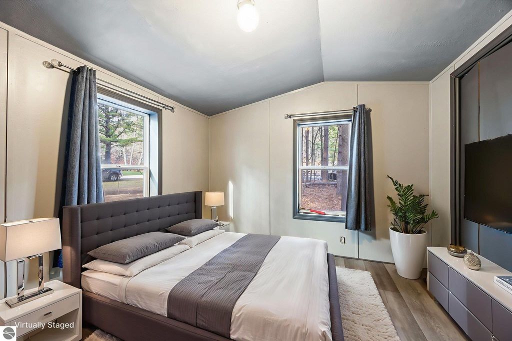 Modern bedroom with a gray upholstered bed, fresh paint, and new flooring, featuring large windows, a bedside table with a lamp, and a decorative plant, showcasing a cozy atmosphere in a move-in ready home at 8401 Independence Avenue, Cadillac, MI.