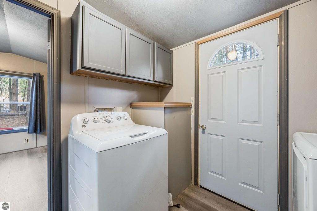 Laundry area with a washing machine, storage cabinets above, and a door leading outside, featuring a view of trees through a nearby window, in a move-in ready home at 8401 Independence Avenue, Cadillac, MI.