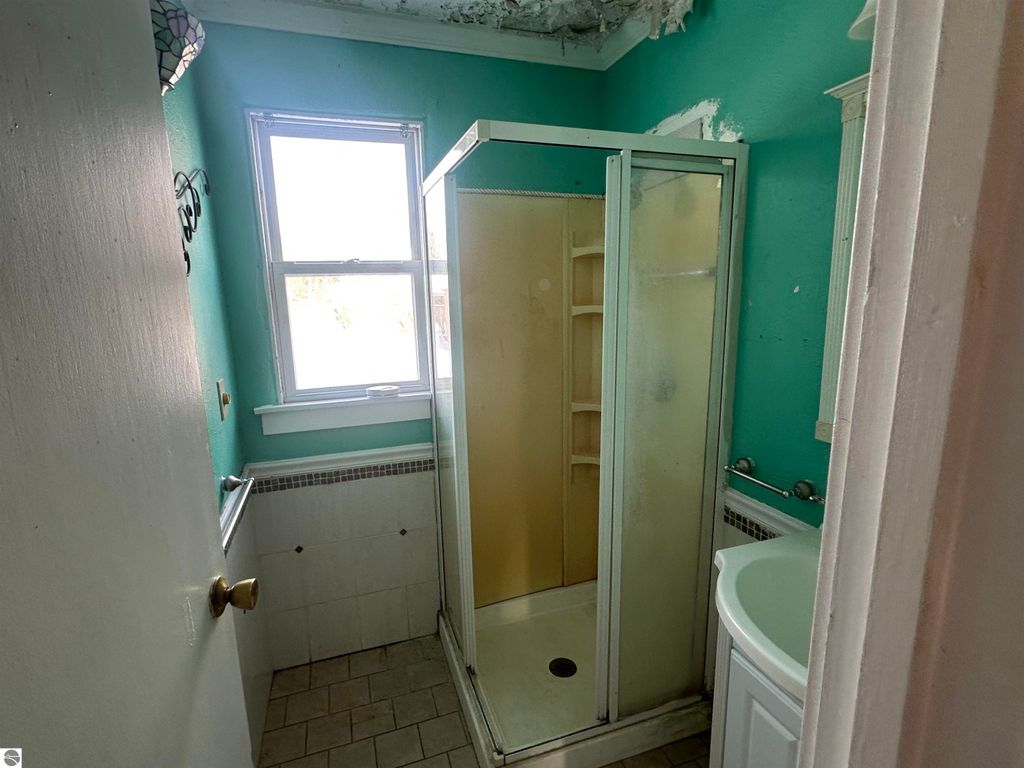 Bathroom interior featuring a turquoise wall, a clear glass shower enclosure, a white vanity sink, and a window providing natural light, showcasing the property's cozy charm at 110 W State Street, Clare, MI.