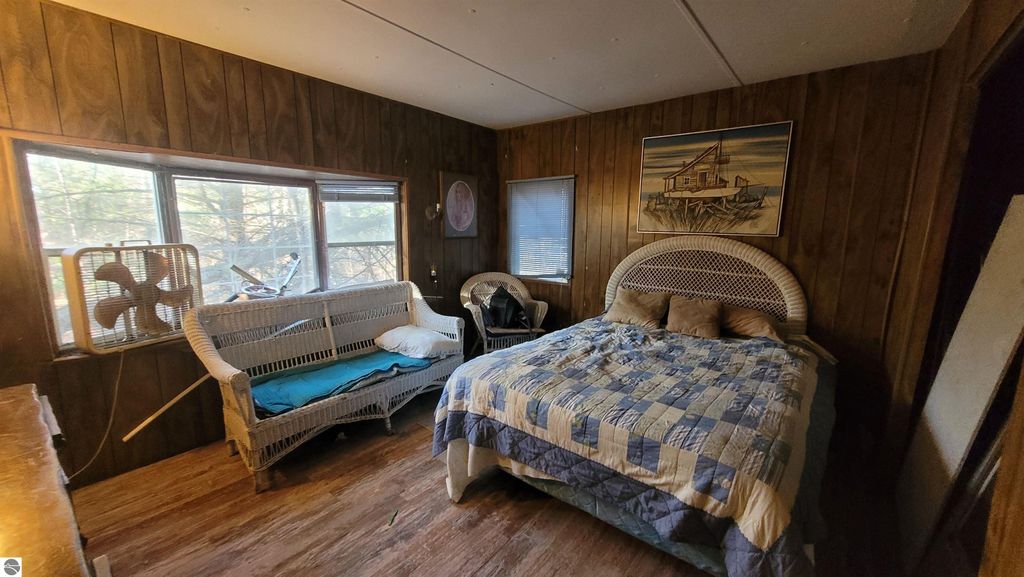 Interior view of a rustic bedroom featuring a bed with a patchwork quilt, a wicker headboard, a wicker sofa, and large windows allowing natural light, suitable for a 2-bedroom mobile home on a 40-acre property in Alden, MI.