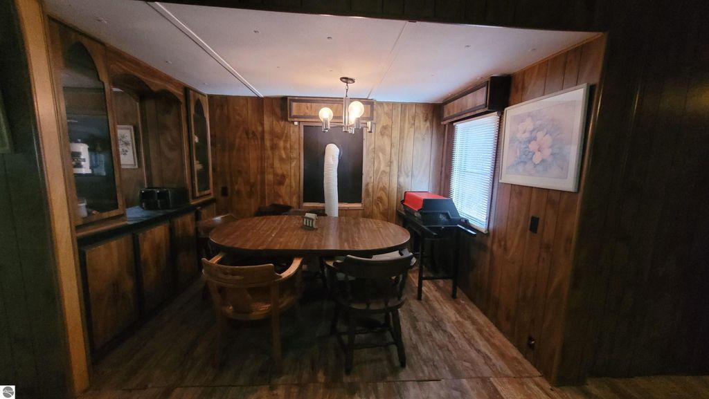 Dining area of a 2-bedroom mobile home, featuring a round wooden table, wooden paneling, and large windows, suitable for renovation or as a hunting camp, part of a 40-acre property in Alden, MI.