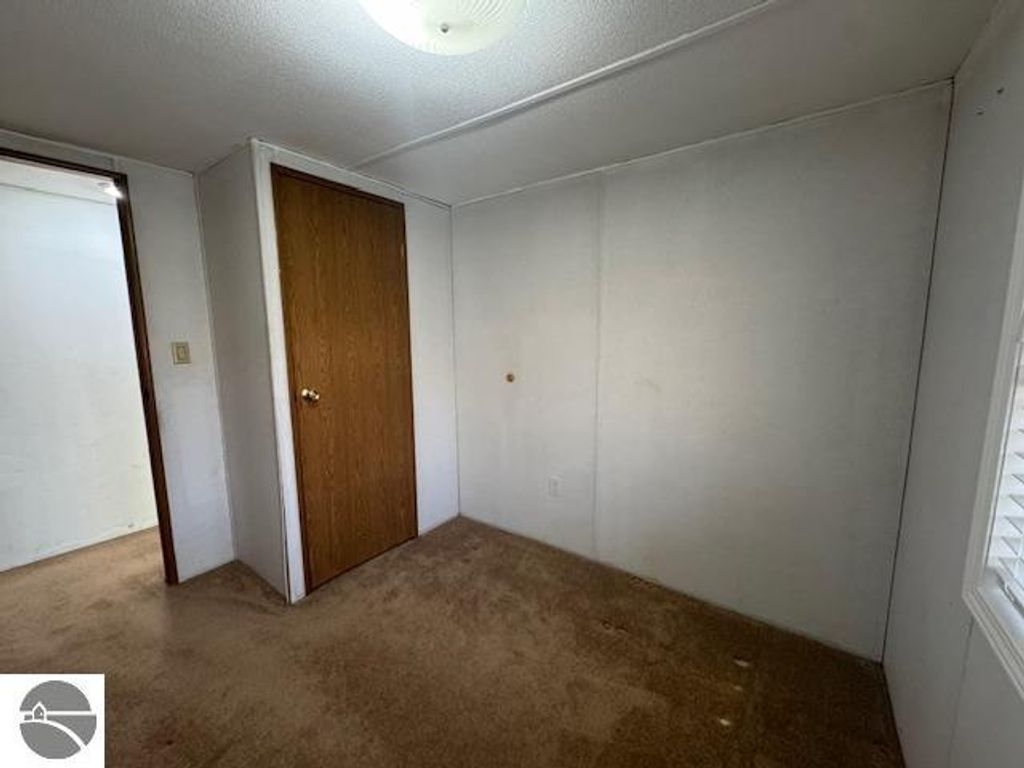 Interior view of a small, empty bedroom featuring beige carpet, a closed wooden door, and a window with blinds, highlighting the space's potential for personalization in the Lake City property listing.